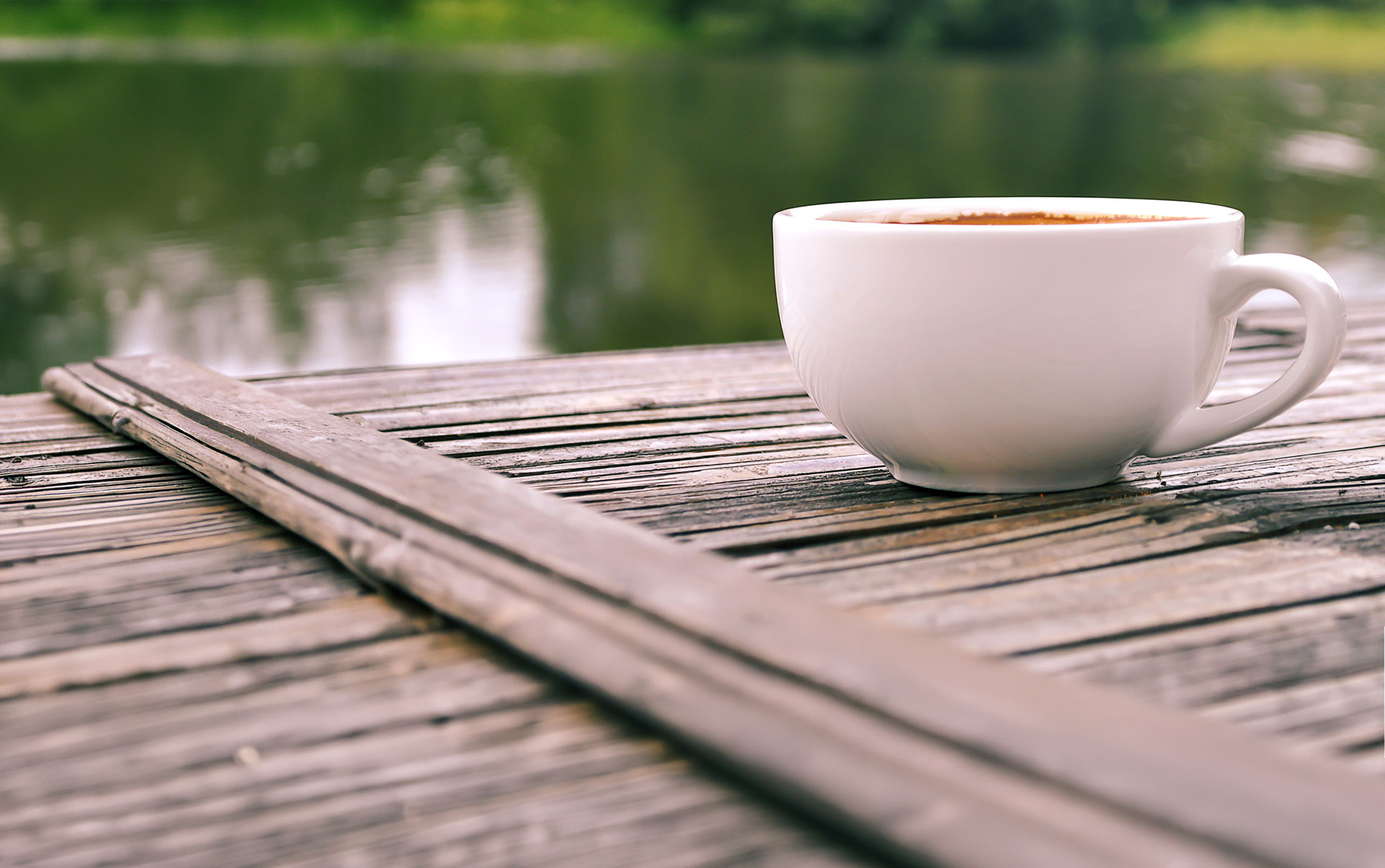 A cup of hot coffee is sat on a wooden table next to the still waters of a lake